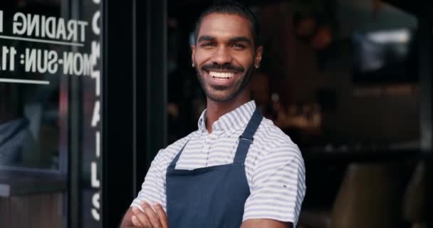 Sorria, braços cruzados e rosto de barista no café para receber com atitude positiva e confiante. Happy, retrato e jovem garçom ou empreendedor do sexo masculino na entrada startup café ou restaurante - Filmagem, Vídeo