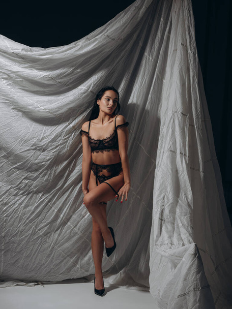 Νεαρή σέξι μελαχρινή γυναίκα ποζάρει με δαντελωτά μαύρα εσώρουχα, ζώνη με τιράντες και ψηλά τακούνια σε σκοτεινό στούντιο σε γκρι φόντο. Στυλ BDSM. - Φωτογραφία, εικόνα