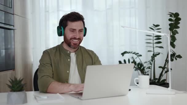 Cheerful man dressed in casual attire shaking head to rhythm of song while typing on personal laptop indoors. Positive male in wireless headphones enjoying remote work at home with favorite music. - Footage, Video
