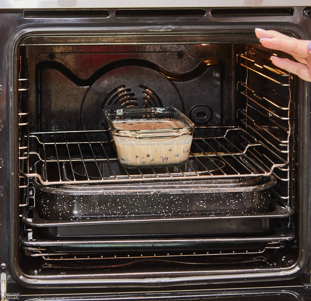 Close-up van een persoon handen samenstellen van een kersentaart in een transparante ovenvorm voor het bakken, met de elektrische oven op de achtergrond. Het bereiden van een heerlijke zelfgemaakte dessert. - Foto, afbeelding