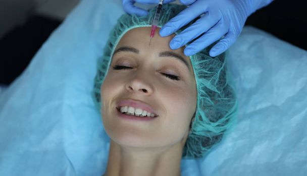 Portrait of smiling lady gets injection of botox in her forehead, cosmetologist using syringe with liquid. Cosmetology, beauty treatment, wellness concept - Foto, afbeelding