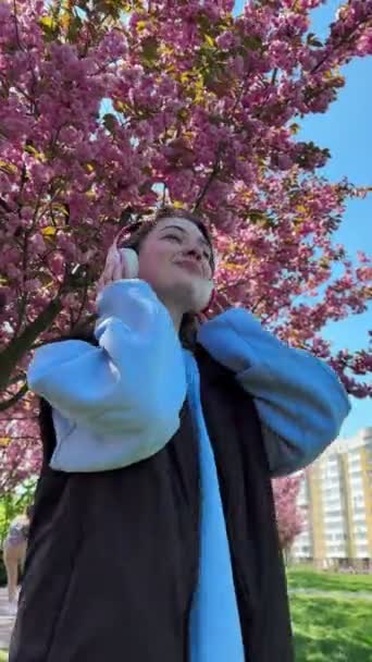 vrouw luisteren muziek in headset bloeiende roze sakura boom op achtergrond - Video