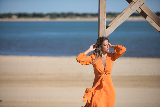 Young and beautiful woman in an orange dress, leaning on a wooden pole, hands on her head, looking at infinity, relaxed and calm, in solitude. Concept beauty, fashion, trend, solitude, peace. - Photo, Image