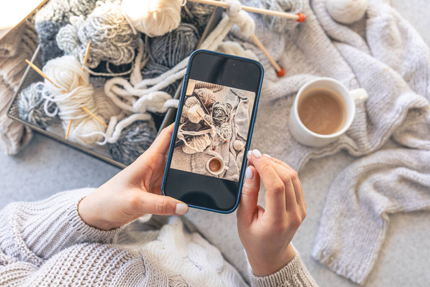 Una donna scatta foto di filati, fili e una tazza di caffè sul suo smartphone. Home hobby concetto. - Foto, immagini