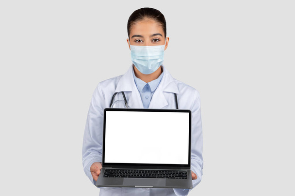 Professioneller europäischer Arzt in weißem Laborkittel und Schutzmaske, präsentiert einen offenen Laptop mit einem leeren Bildschirm für medizinische Informationen, isoliert auf grauem Studiohintergrund - Foto, Bild
