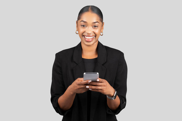 Femme d'affaires afro-américaine souriante utilisant un smartphone, engagée dans la communication moderne, représentant l'accessibilité et la connectivité dans une tenue professionnelle sur fond gris, studio - Photo, image