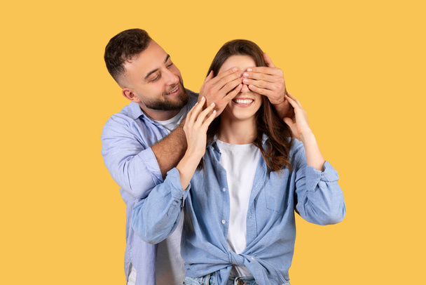 Zabawny młody mężczyzna zaskakuje śmiejącą się kobietę zakrywając jej oczy dłońmi, dzieląc radosną chwilę razem na żółtym tle - Zdjęcie, obraz