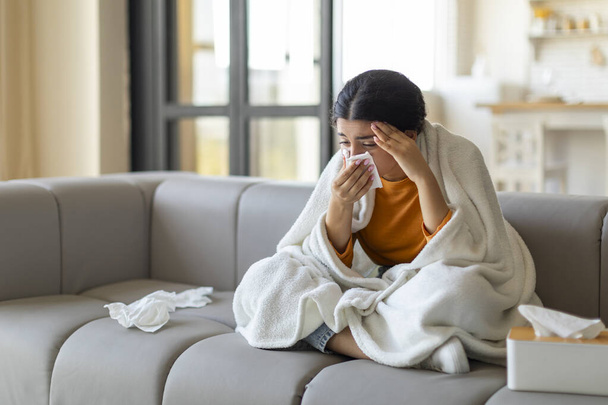 Indian woman feeling ill, sneezing into tissue, sick eastern lady wrapped in blanket sitting on couch at home, upset female suffering seasonal flu or cold, having influenza symptoms, free space - Photo, Image