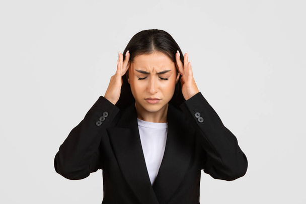 Mujer de negocios europea triste estresada en traje negro tocando sus sienes, mostrando signos de dolor de cabeza o concentración intensa, sobre fondo gris claro - Foto, imagen