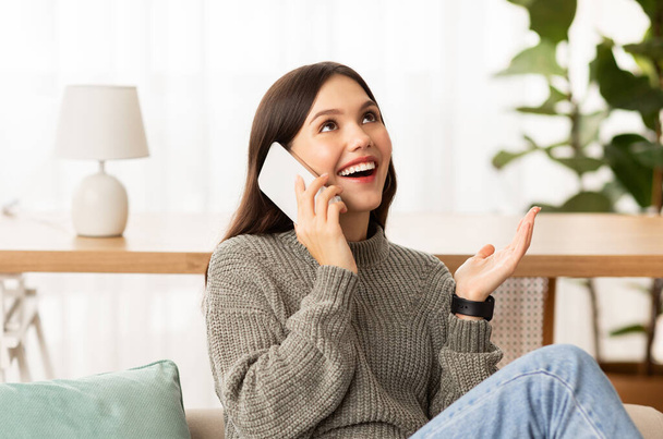 Communication concept. Emotional beautiful brunette young woman wearing casual clothing sitting on couch in cozy living room interior at home, talking on phone, laughing and gesturing - Photo, Image