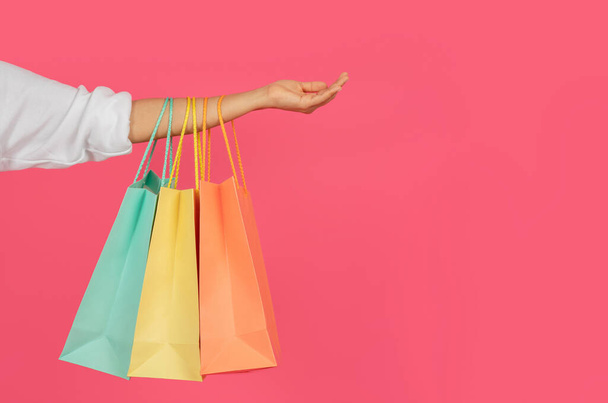 Kolorowe torby na zakupy powieszone na kobiecej dłoni na różowym tle pracowni, nierozpoznawalna zakupoholiczka przewożąca zakupy, ciesząca się sezonową sprzedażą i rabatami, ucięte ujęcie z przestrzenią do kopiowania - Zdjęcie, obraz