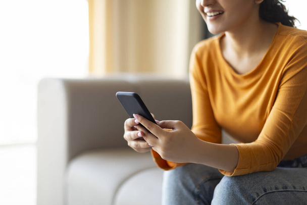 Happy Young Woman SMS-y na smartfonie podczas relaksu na kanapie w domu, Millennial Kobieta za pomocą telefonu komórkowego do komunikacji online, siedzi na wygodnej kanapie w salonie, przycięte - Zdjęcie, obraz