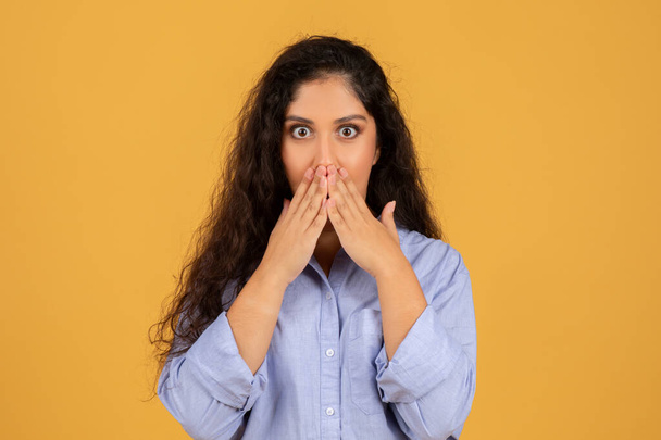 Startled young middle eastern woman with voluminous curly hair covering her mouth with hands, eyes wide in shock, wearing a light blue shirt against a mustard yellow background - Фото, зображення