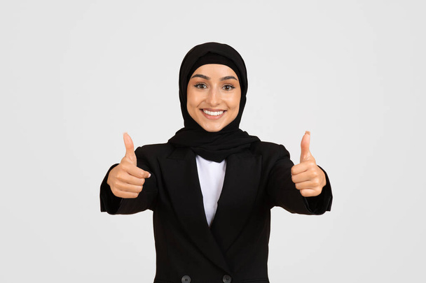 ハッピーイスラム教のヨーロッパの実業家は,両手で親指をあげて笑顔を浮かべ,黒いスーツとヒジャブを着て承認と成功を象徴しています - 写真・画像