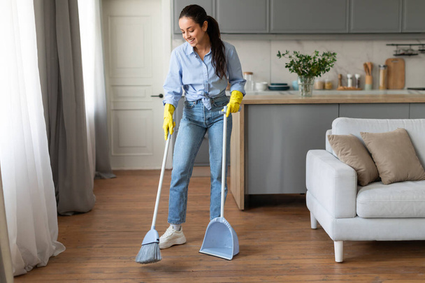 Focused young woman in casual outfit and yellow gloves sweeping the floor with broom and holding dustpan in bright, cozy home interior - Photo, Image