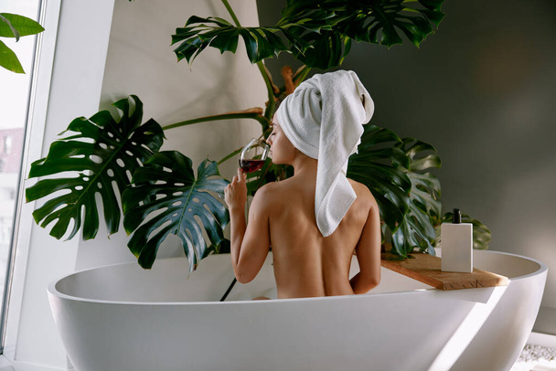 Schöne Frau beim Baden im Badezimmer mit einem Glas Wein. Wellness-Behandlung, Haut- und Körperbehandlung zu Hause, Entspannung und Wohlbefinden - Foto, Bild