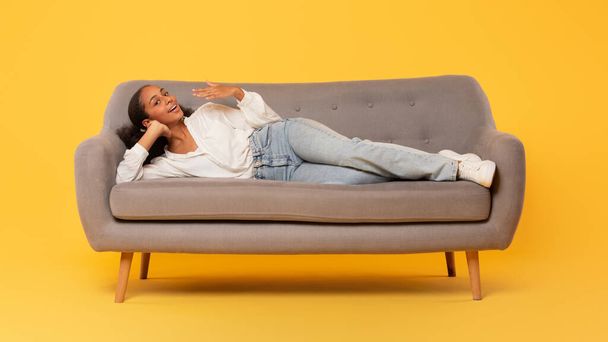 Повна довжина розслабленої чорної дівчини-підлітка в повсякденному вбранні лежить на зручному дивані в студії на жовтому тлі. Пані-підліток насолоджується вільним часом і розслабленням. Панорама - Фото, зображення