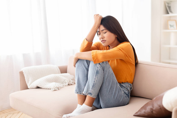 Upset young asian woman sitting on couch with legs up, touching her head, suffering from migraine during pms or period. Sad chinese lady feeling lonely, depressed, frustrated - Photo, Image