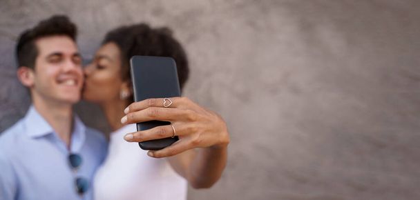 Coppia multiculturale che condivide un momento romantico mentre si fa un selfie con uno smartphone su uno sfondo strutturato. Concentrati sul telefono, facce sfocate. - Foto, immagini
