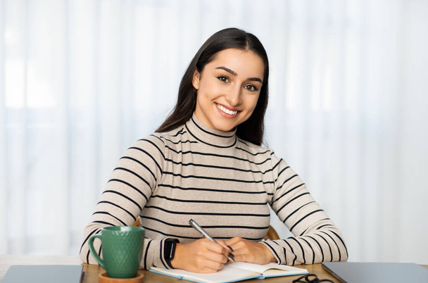 Mulher do Oriente Médio jovem confiante com um sorriso encantador sentado em casa, escrevendo em seu caderno com uma caneca verde e tablet na mesa, exalando produtividade e contentamento - Foto, Imagem