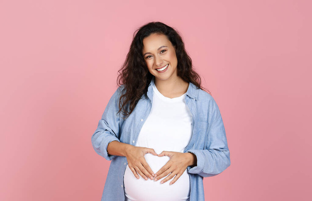 Positive expecting millennial European woman hold hands in heart shape on her big tummy, smiling at camera. Happy young pregnant lady posing isolated on pink background, copy space - Photo, Image