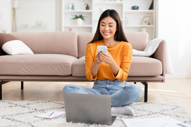 Pěkně usměvavá mladá asijská žena sedící na podlaze s laptopem, pomocí smartphonu, čínská dáma podnikatel pomocí obchodní mobilní aplikace, práce z domova, kontrola e-mailu, kopírovací prostor - Fotografie, Obrázek