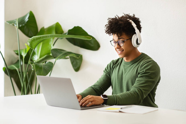 Веселый молодой африканский американец проводит онлайн-встречу с коллегами, работающими из кафе или домашнего офиса, сидящими за столом, используя компьютерные ноутбуки и беспроводные наушники, телекоммуникации - Фото, изображение