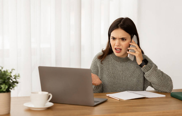 Wściekła młoda bizneswoman odbyć rozmowę telefoniczną z asystentem lub partnerem biznesowym, siedzi przy biurku w biurze, patrząc na ekranie laptopa, gestykulując - Zdjęcie, obraz