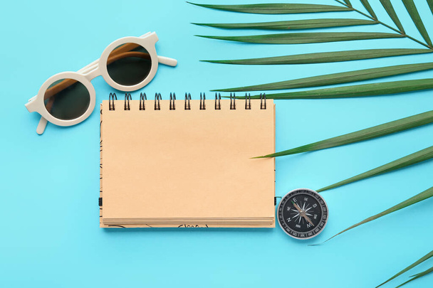 Composición con cuaderno en blanco, hoja de palma, gafas de sol elegantes y brújula sobre fondo azul - Foto, imagen