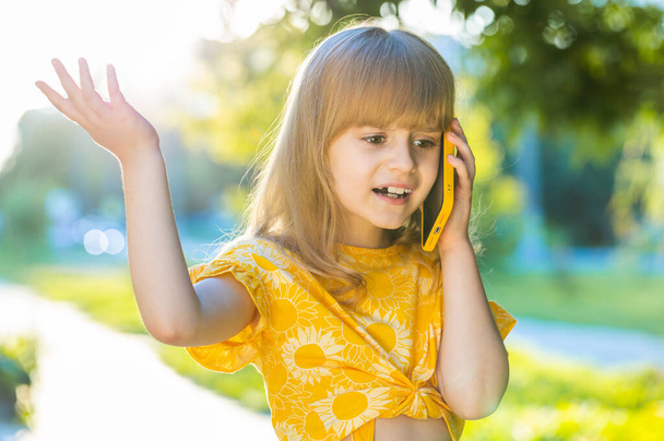 Грустный ребенок-подросток имеет дистанционный разговор общаться разговаривая по смартфону. Красивая блондинка дочь разговаривает с матерью по телефону сплетни стоя на городской закат парк-стрит - Фото, изображение