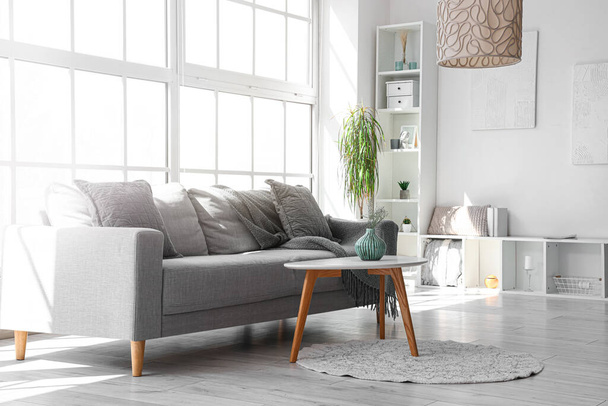Accogliente divano grigio con cuscini e tavolino in luminoso soggiorno vicino a una grande finestra - Foto, immagini