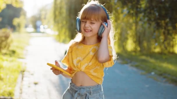 Happy overjoyed pre teen dítě v bezdrátových sluchátkách výběru, poslouchat oblíbené energické disco rock n roll hudby v tanci smartphone. Blondýny dívka chůze na město západ parku ulici, venku - Záběry, video
