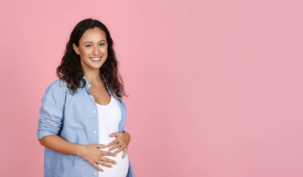 Беременность, материнство. Счастливая беременная женщина за 30, одета в повседневную одежду, позирует изолированно на розовом фоне студии, держит за руки свой большой живот и улыбается в камеру, копирует пространство, панораму - Фото, изображение