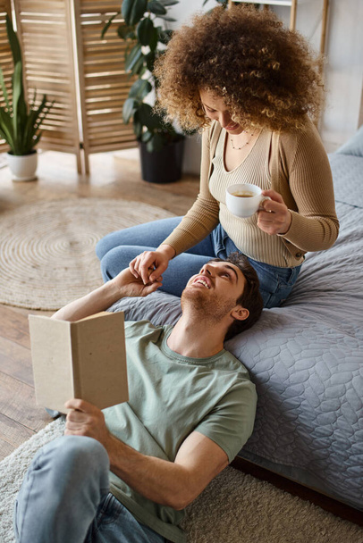 Hangulatos beszélgetés kávé és könyv az ágyban, göndör nő és barna férfi élvezi egymás társaságát - Fotó, kép