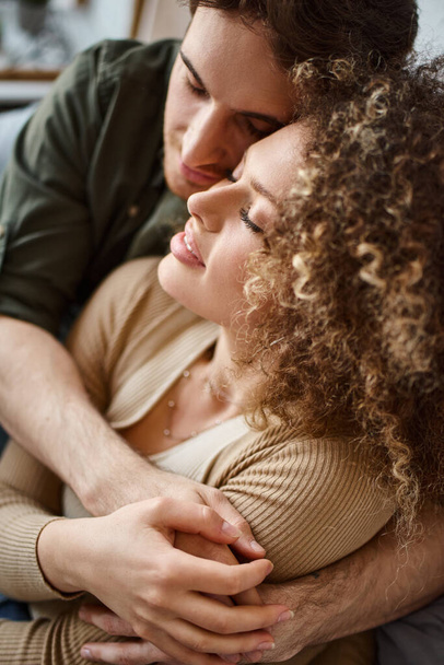 Κοντινή φωτογραφία με μια σγουρή νεαρή γυναίκα και έναν μελαχρινό άντρα να μοιράζονται μια εγκάρδια αγκαλιά στην κρεβατοκάμαρα - Φωτογραφία, εικόνα