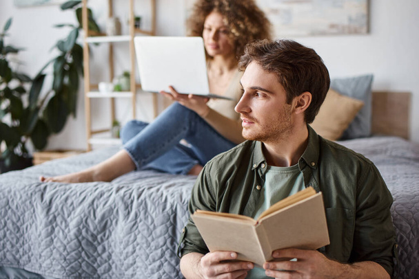 Portrait d'homme tenant livre tandis que sa petite amie utilise un ordinateur portable couché sur le lit, temps libre - Photo, image
