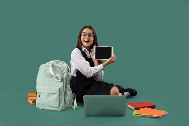 Little schoolgirl with tablet computer, laptop and backpack on green background - Photo, Image