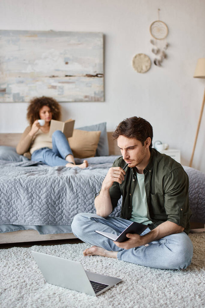 Paar verbringt Zeit zu Hause. Frau liest Buch im Bett und Mann macht sich Notizen auf dem Fußboden - Foto, Bild