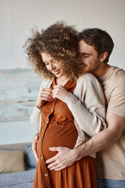 Прекрасный счастливый мужчина обнимает беременную жену сзади, стоя дома и ожидая ребенка. - Фото, изображение