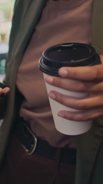 Close-up vertical tilting shot of frowning young African American female entrepreneur or office worker in blazer and t-shirt holding takeaway coffee and typing message or email on smartphone - Footage, Video