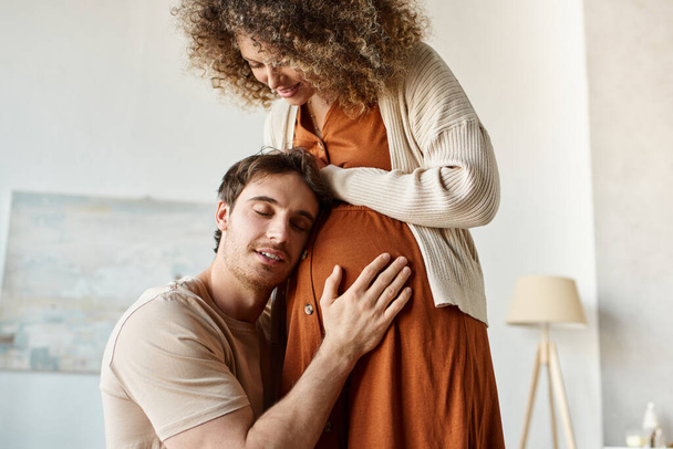 Jóképű, mosolygós, boldog férfi hallgatja, ahogy a baba ölelgeti a feleségét, megérinti a hasát. - Fotó, kép