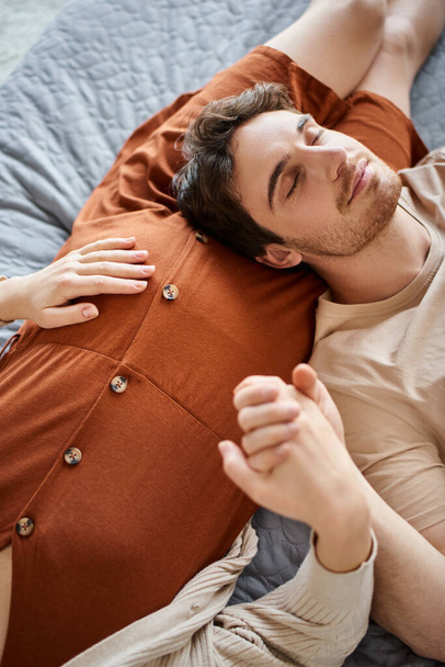 Έγκυος γυναίκα με σύζυγο ξαπλωμένη στο κρεβάτι, άντρας αναπαύεται κοντά στην κοιλιά της γυναίκας του και χαμογελάει - Φωτογραφία, εικόνα