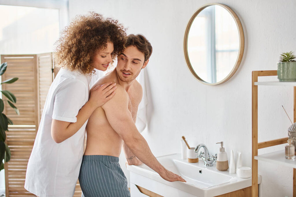 Kıvırcık genç kadın ve esmer adamın banyoda birbirine sarılıp gülümsemesinin yan görüntüsü. - Fotoğraf, Görsel