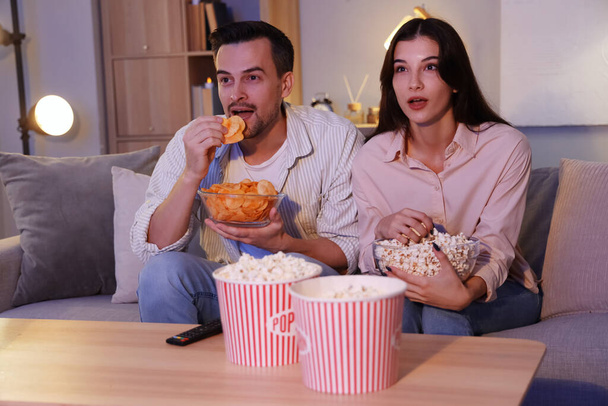 Młoda para z chipsami ziemniaczanymi i popcornem ogląda wieczorem telewizję w domu - Zdjęcie, obraz