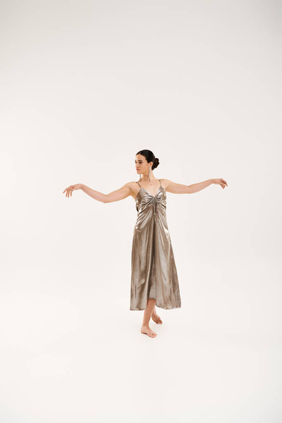 Μια νεαρή γυναίκα με ένα ασημένιο φόρεμα χορεύει με χάρη, εκφράζοντας την κομψότητα και την κίνηση σε ένα στούντιο σε ένα λευκό φόντο. - Φωτογραφία, εικόνα