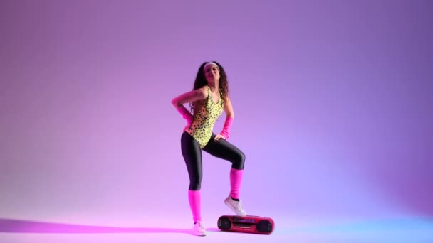 Lockige 80er-Jahre-Frau groovt in lebendiger Retro-Kleidung vor violettem Hintergrund und tanzt mit einem Kassettenspieler auf dem Boden. Ihre lebhaften Bewegungen zeigen eine lustige und nostalgische Ära. - Filmmaterial, Video