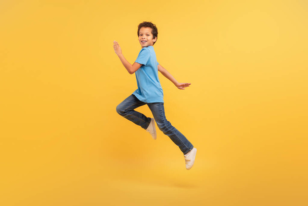 Ενεργητικό μαύρο αγόρι που συλλαμβάνεται στη μέση του αγώνα με χαρούμενη έκφραση, αθλητικό μπλε t-shirt και τζιν, με φόντο το ζωηρό κίτρινο φόντο - Φωτογραφία, εικόνα