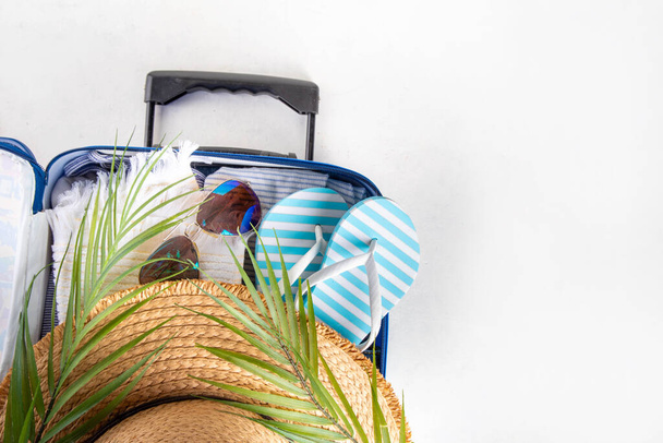 Tatil, tatil, seyahat ve seyahat konsepti. Elbiseli bavul, plaj aksesuarları, şapka, güneş gözlüğü, parmak arası terlik, beyaz arka planda tropikal yapraklar fotokopi alanı - Fotoğraf, Görsel