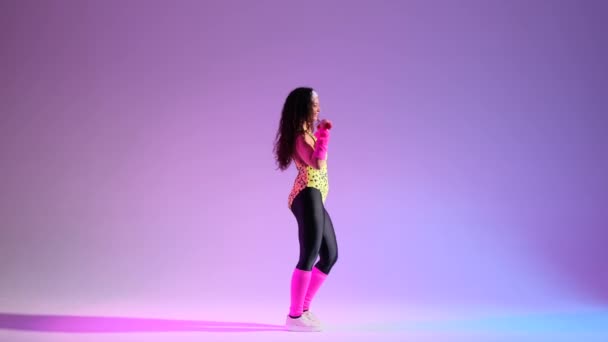 Girl in a 80s retro outfit engages in energetic aerobics with dumbbells against a vivid purple background. Mezcla de fusión dinámica de fitness y estilo retro. - Metraje, vídeo
