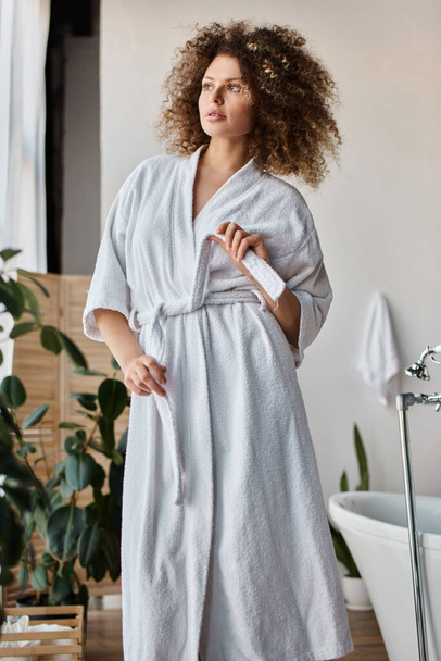 Schöne lockige junge Frau trägt weiße Robe im Badezimmer binden den Gürtel und schaut weg - Foto, Bild